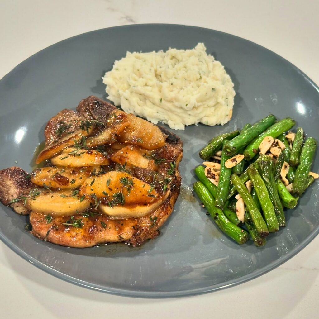 Apple-Cider Glazed Pork Chops served with creamy garlic mashed potatoes and sautéed green beans topped with toasted almonds on a white marble countertop by Reynaldo’s Cocina.