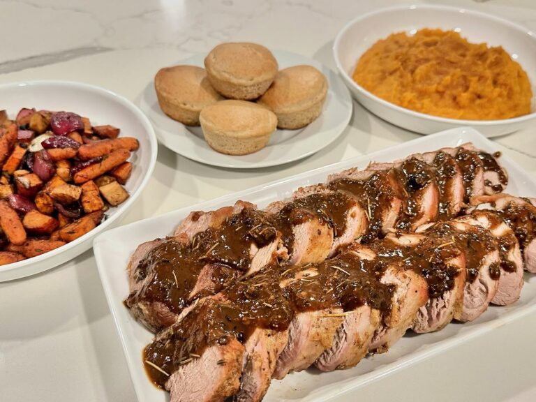 Sliced oven-roasted pork tenderloin finished with homemade apple cider glaze and served with maple whipped sweet potatoes, roasted root vegetables, and homemade apple cider muffins on a white marble countertop.