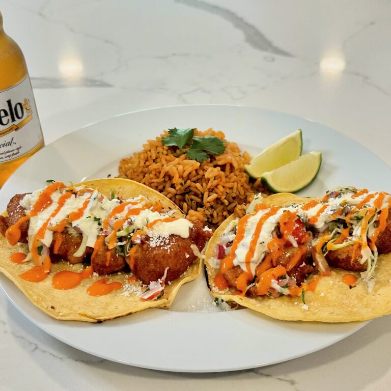 Beer-battered shrimp tacos topped with cabbage, crema, hot sauce, cotija cheese, and served with Mexican rice and lime wedges.