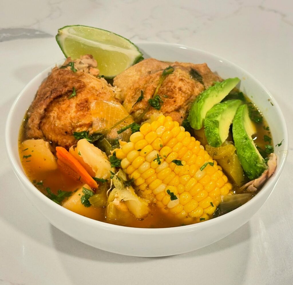 Caldo de Pollo Reynaldos Cocina Homemade Caldo de Pollo chicken soup with corn, potatoes, carrots, avocado, and lime served in a white bowl from Reynaldo’s Cocina
