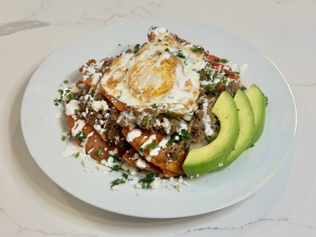 Homemade chilaquiles rojos made with crispy fried tortilla chips tossed in guajillo sauce and topped with shredded beef, fried egg, queso fresco, crema, cilantro, and avocado.