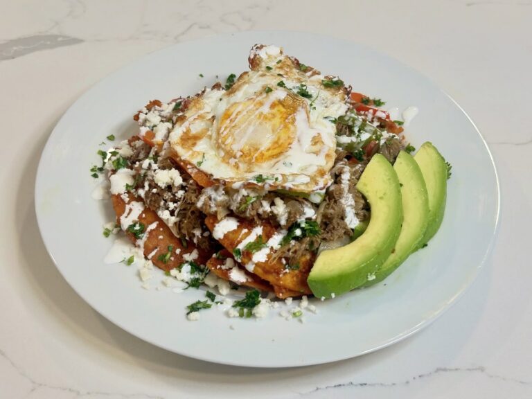 Homemade chilaquiles rojos made with crispy fried tortilla chips tossed in guajillo sauce and topped with shredded beef, fried egg, queso fresco, crema, cilantro, and avocado.