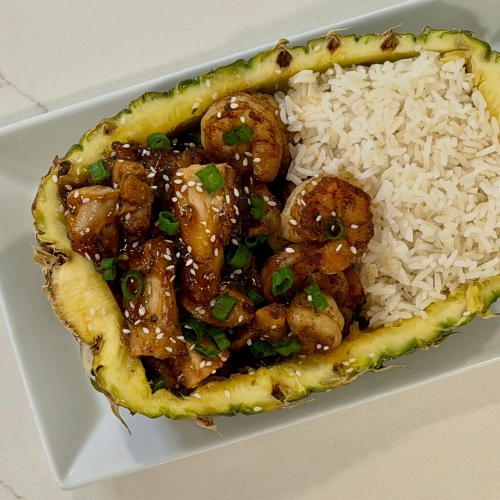 Homemade honey sesame chicken and shrimp served inside a fresh pineapple bowl with jasmine rice, topped with sesame seeds and green onions.