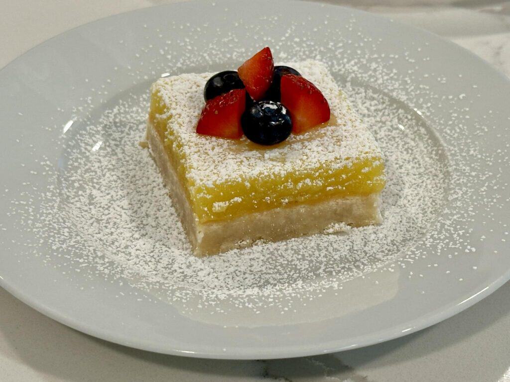 Homemade lemon bars topped with strawberries, blueberries, and powdered sugar on a white plate.