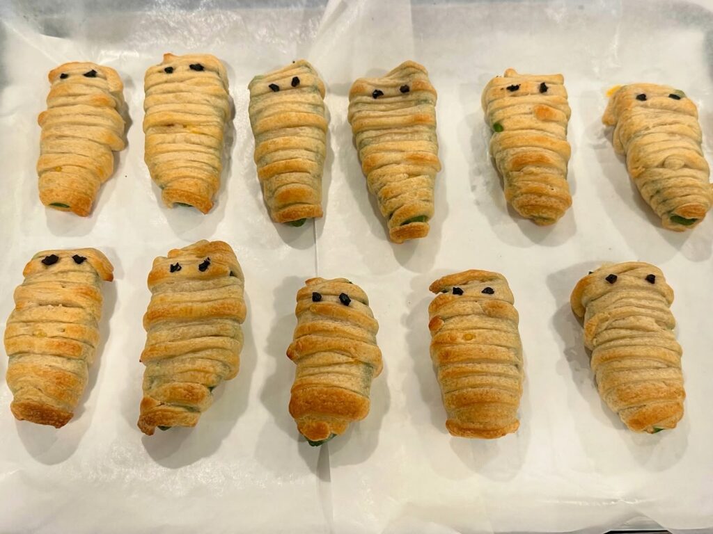 Homemade Mummy Jalapeño Poppers stuffed with cream cheese, cheddar cheese, and bacon then wrapped in crescent dough strips with black olive eyes.