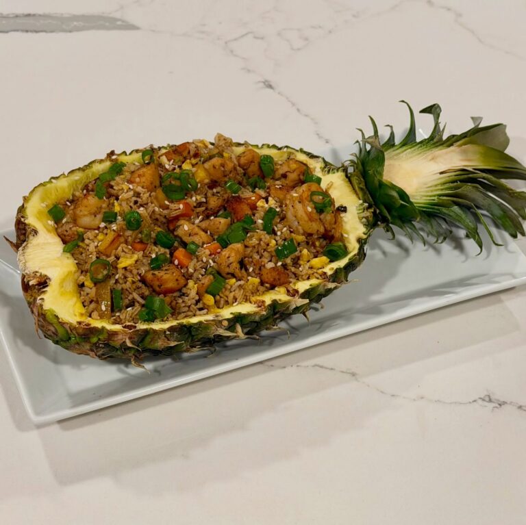 Homemade pineapple chicken and shrimp fried rice served inside a caramelized pineapple shell with garlic, ginger, eggs, green onion, pineapple chunks, and honey sesame sauce.