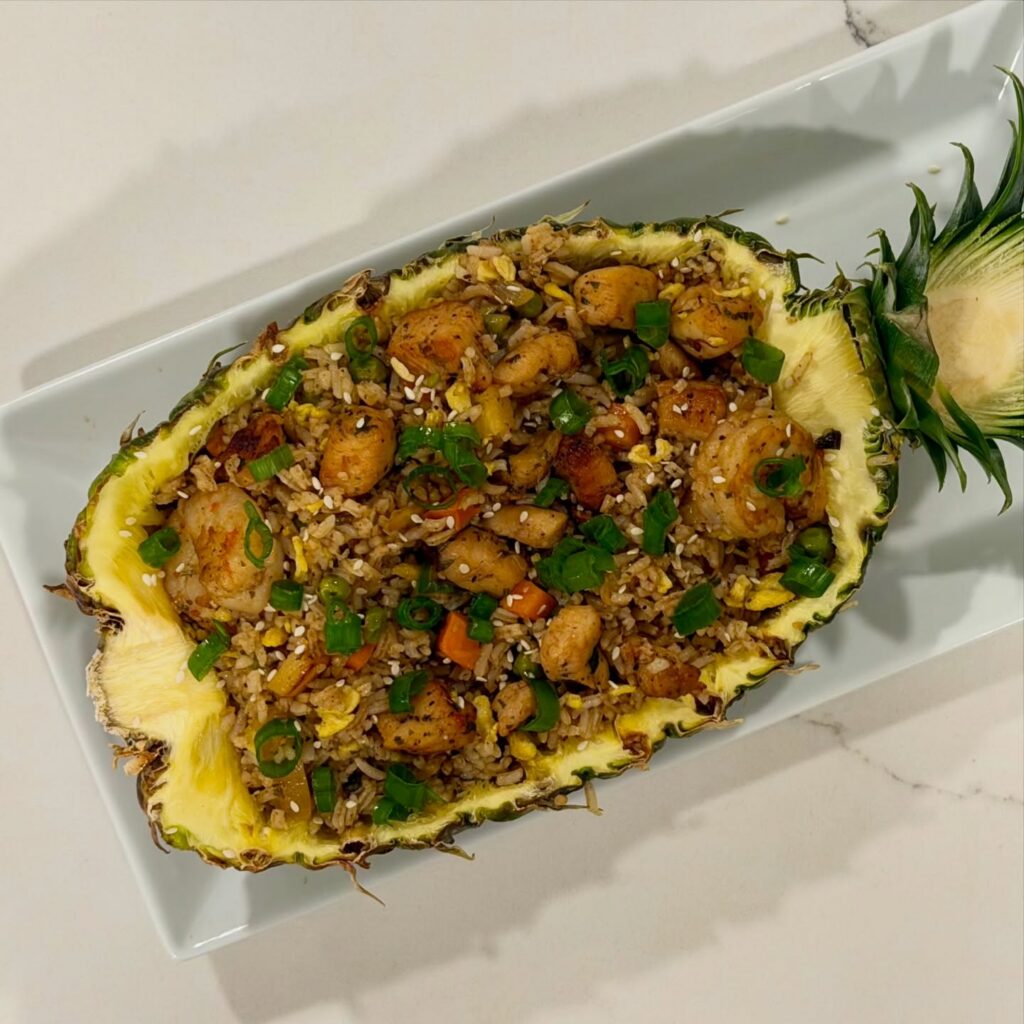 Homemade pineapple chicken and shrimp fried rice served inside a caramelized pineapple shell with garlic, ginger, eggs, green onion, pineapple chunks, and honey sesame sauce.