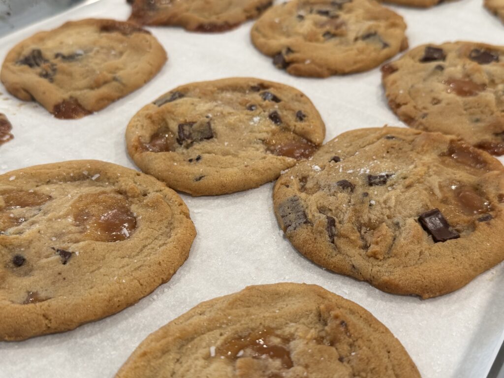Fresh baked salted caramel chocolate chip cookies cooling on parchment paper with melted caramel and chocolate chunks.