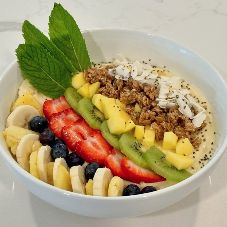 Tropical smoothie bowl topped with kiwi, strawberries, blueberries, pineapple, banana, mango, granola, coconut, chia seeds, and fresh mint.