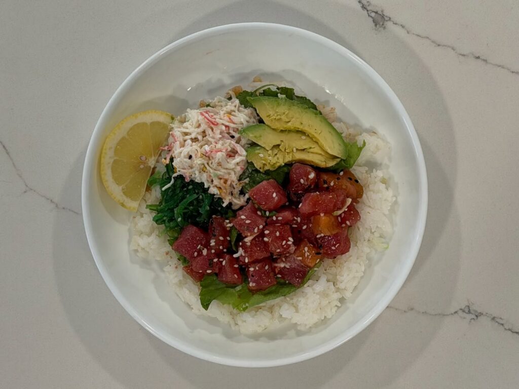 Homemade tuna and salmon poke bowl with sushi rice, marinated ahi tuna, salmon, crab salad, wakame seaweed salad, avocado, sesame seeds, and fresh lemon.