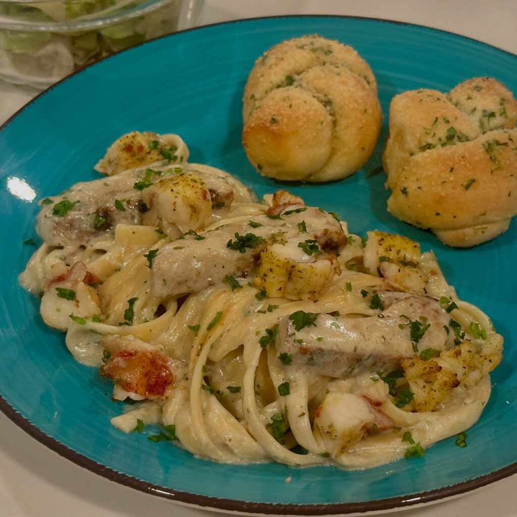Plate of homemade Surf and Turf Fettuccine Alfredo with grilled chicken, buttery lobster, creamy Parmesan Alfredo sauce, fresh parsley, garlic knots, and side salad.